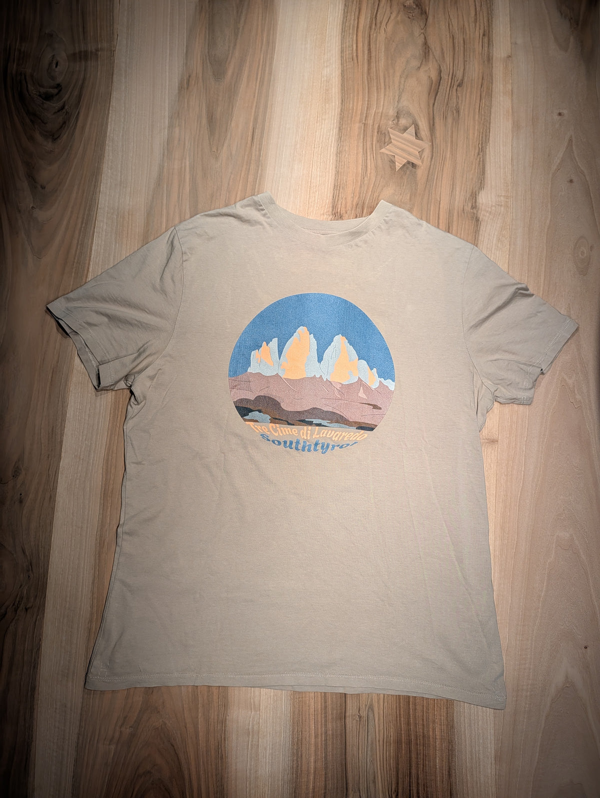 Tre Cime di Lavaredo t-shirt in heather dust, flat lay on wood – close-up of the three peaks design